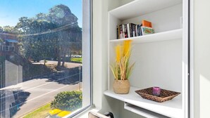 A room with a window overlooking a street, a bookshelf with books, a woven basket with a candle, and a small plant. - Bayview Towers 6, 15 Victoria Pde - stunning water views (Nelson Bay)