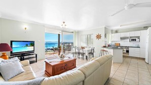 A modern living room with a TV, sofa, and dining area. There's a kitchen with white cabinets and a view of the ocean. - Bayview Towers 6, 15 Victoria Pde - stunning water views (Nelson Bay)