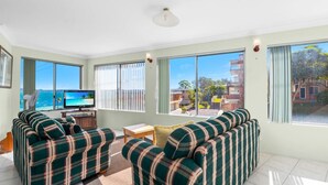 A living room with a sofa set, a TV, and large windows with a view of the sea and buildings. - Bayview Towers 6, 15 Victoria Pde - stunning water views (Nelson Bay)