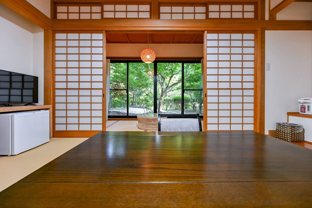 Een traditionele Japanse kamer met tatami vloerbedekking, een televisie en uitzicht op een tuin door een shoji scherm.