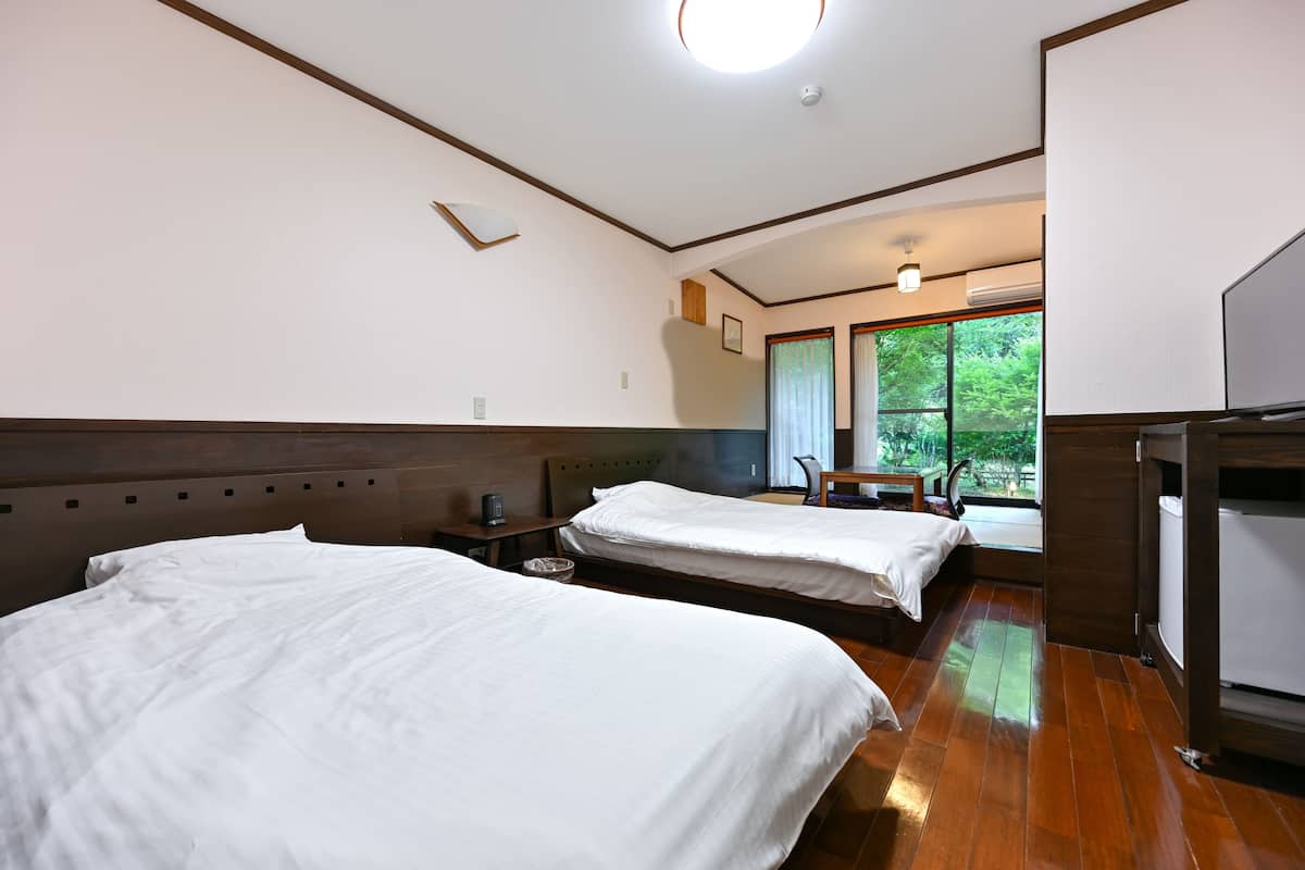 Een hotelkamer met twee bedden, een houten hoofdbord, een bureau met stoel, een raam met uitzicht op groen en een televisie.