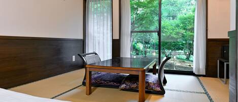 Ein Zimmer mit einem Bett, einem Tisch und Blick auf Grün hinter großen Fenstern.