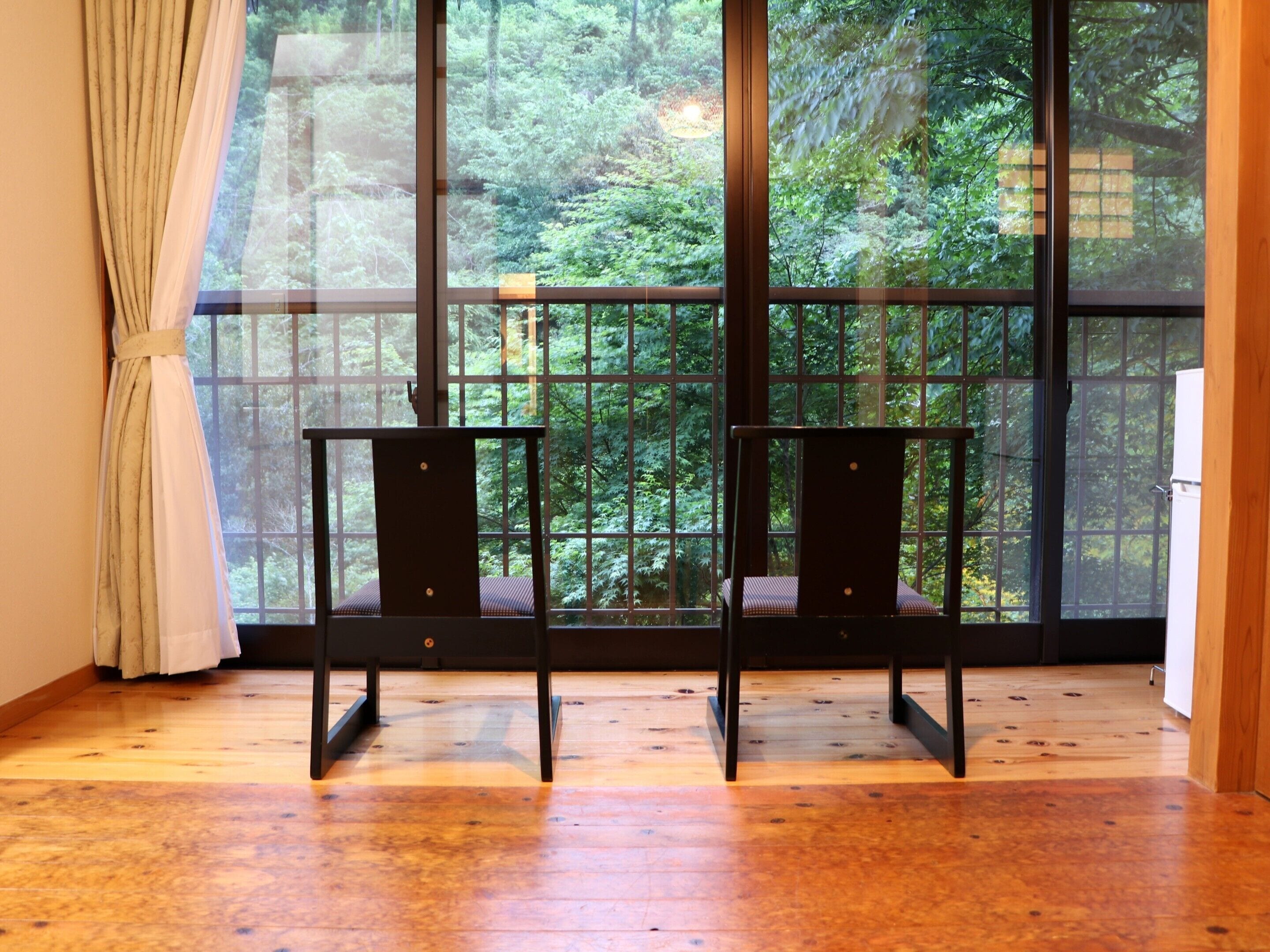 Una habitación con un ventanal que ofrece vistas a un bosque. Hay dos sillas negras y una pequeña mesa en la habitación.