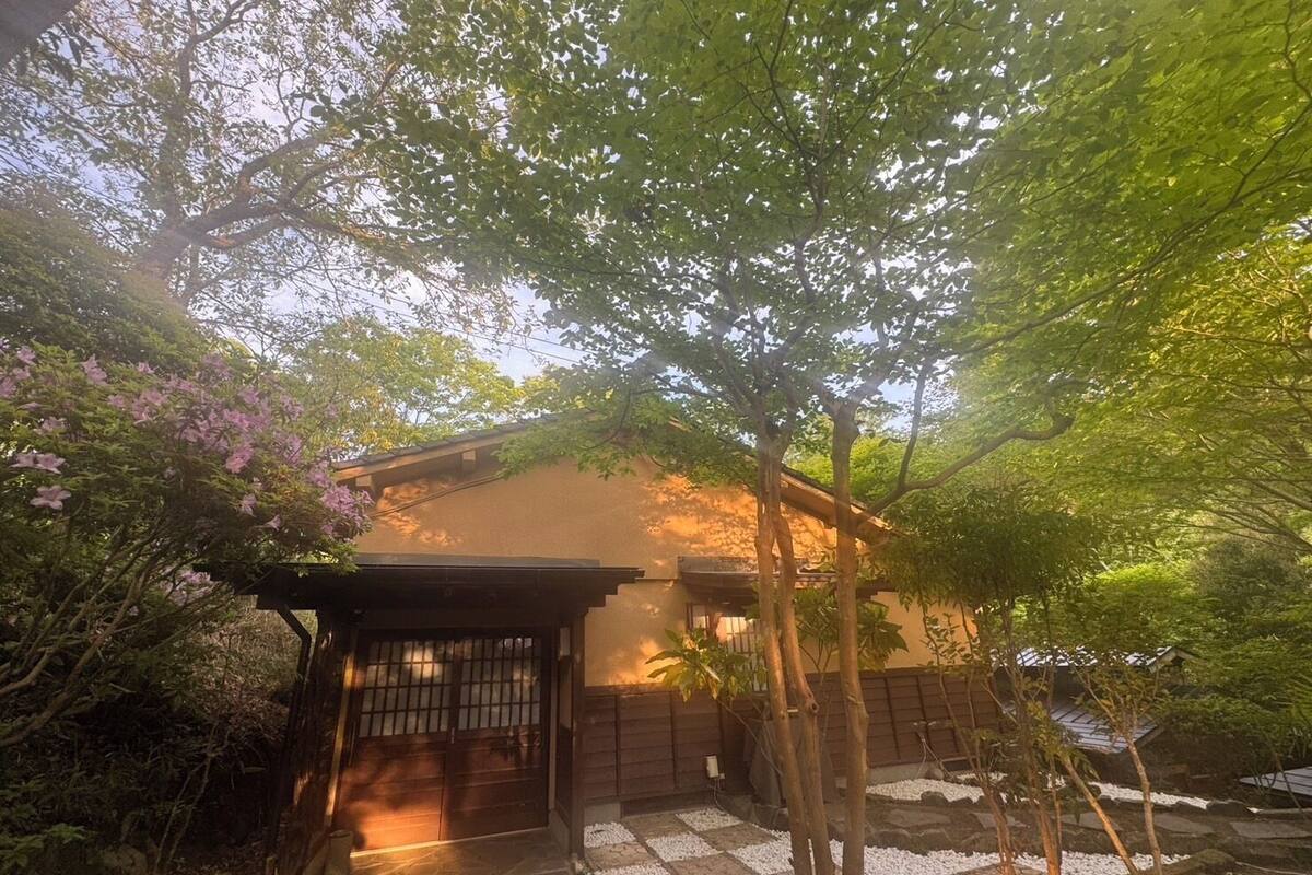 Een traditioneel Japans huis met een houten poort, omgeven door weelderige begroeiing en bloeiende bomen.