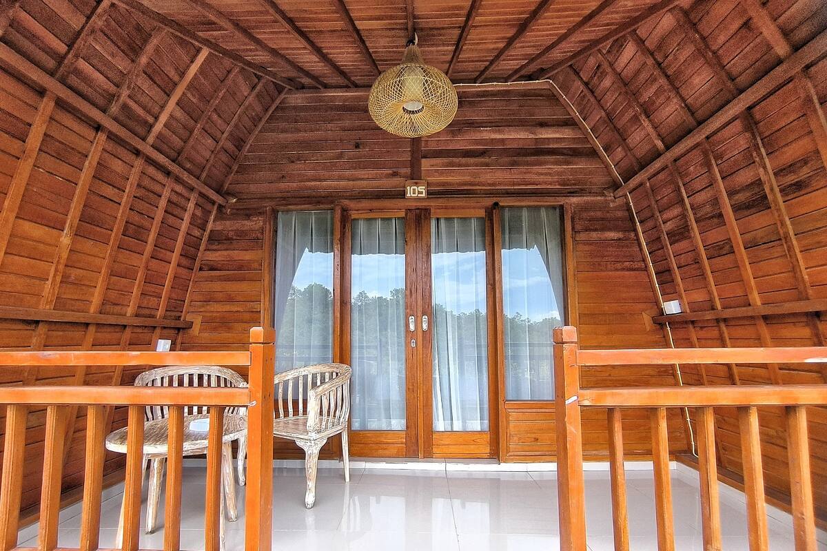 An attic room with a wooden ceiling, a large window with curtains, and a wooden railing.