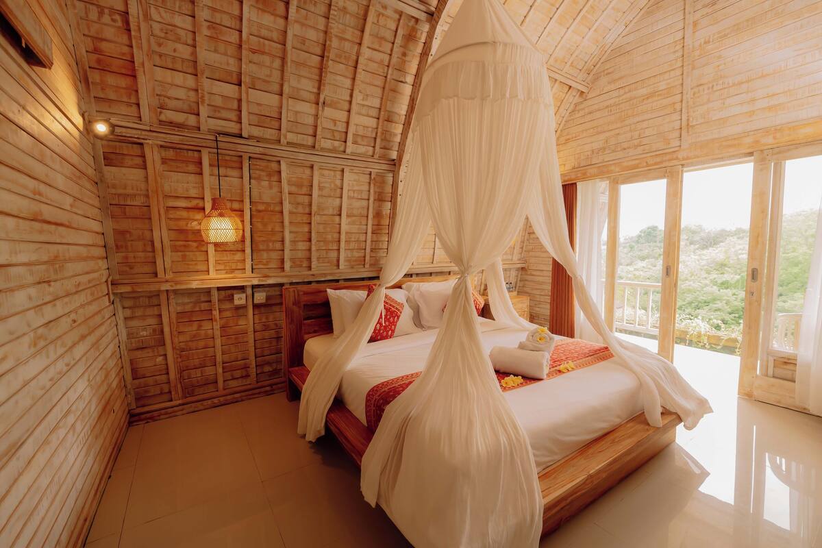 A wooden room with a bed, a hanging light fixture, and a view through large windows.