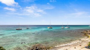 A beach with clear turquoise water, several sailboats anchored, and a rocky shoreline.