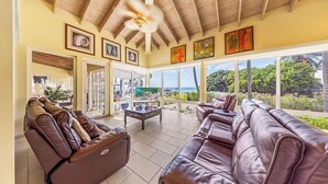 A spacious living area with leather sofas, a ceiling fan, and large windows offering a view of the outdoors.