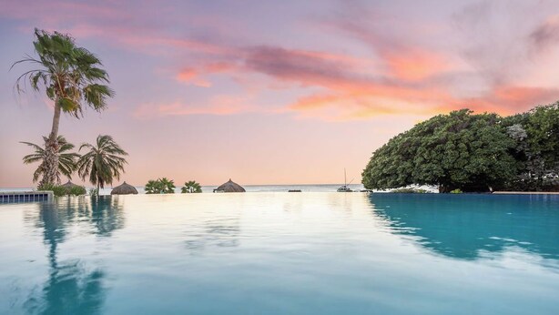 A clear view of a swimming pool with a sunset in the background, surrounded by palm trees and a small island with huts.
