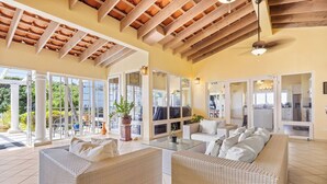 A spacious living area with wicker furniture, large windows, and a wooden ceiling.