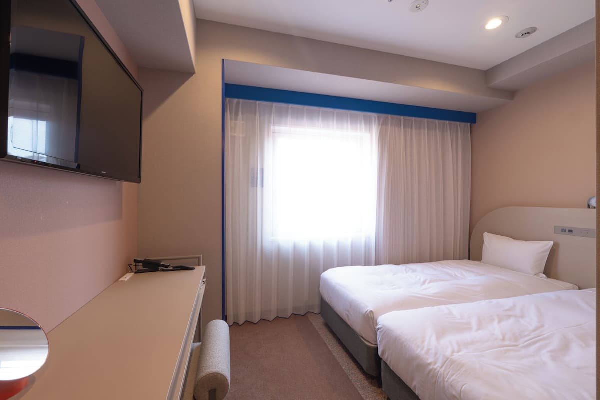 A hotel room with two beds, a flat-screen TV mounted on the wall, a window with sheer curtains, and a small desk with a lamp.