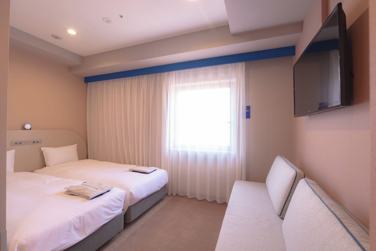 A hotel room with two beds, a sofa, a flat-screen TV, and a window with sheer curtains.
