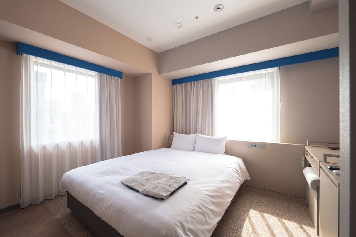 A hotel room with a large bed, two windows with blue curtains, a desk with a chair, and a small bedside table.