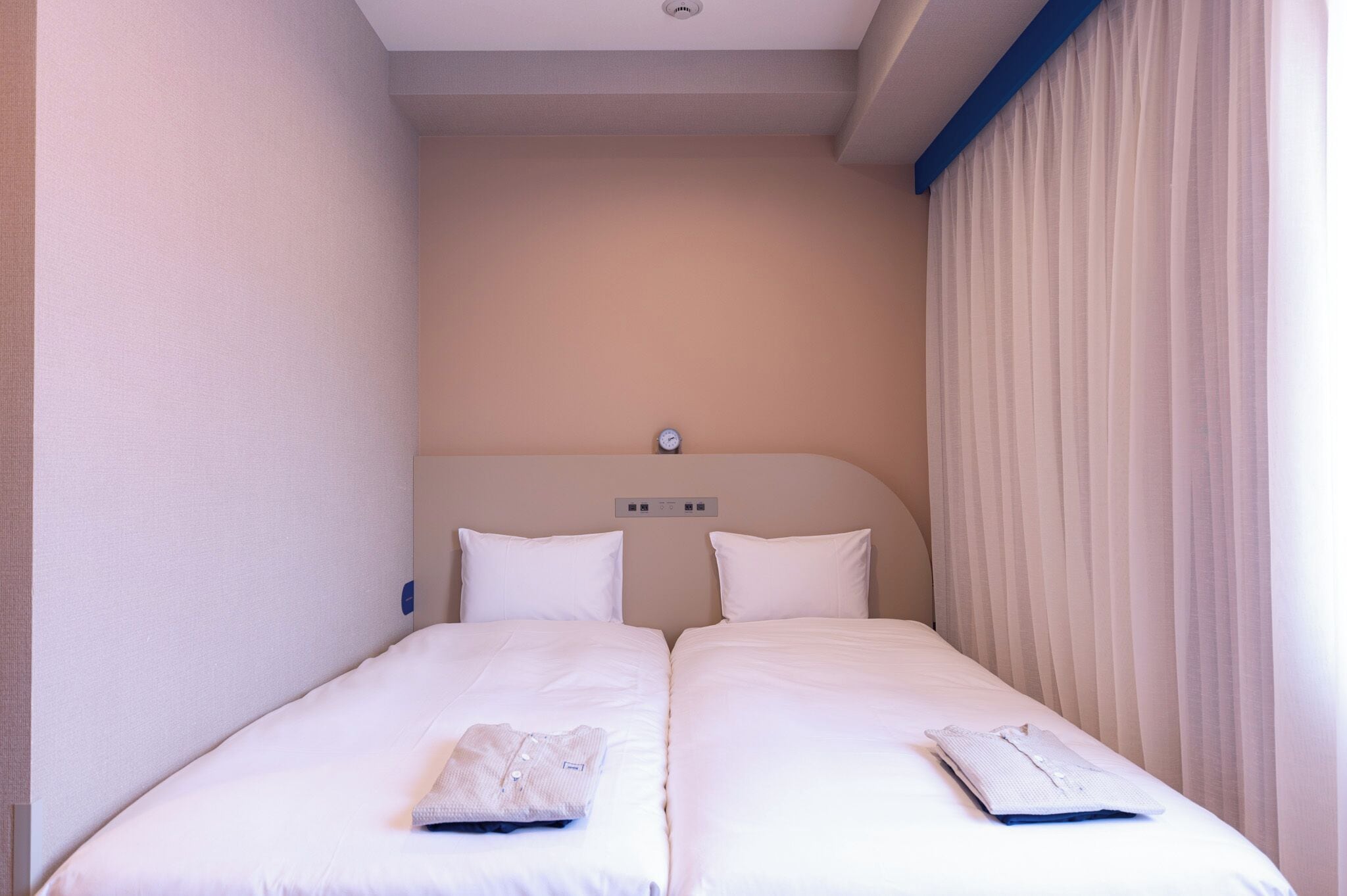 Een hotelkamer met twee enkele bedden, een hoofdeinde met een klok, en een raam met gordijnen.