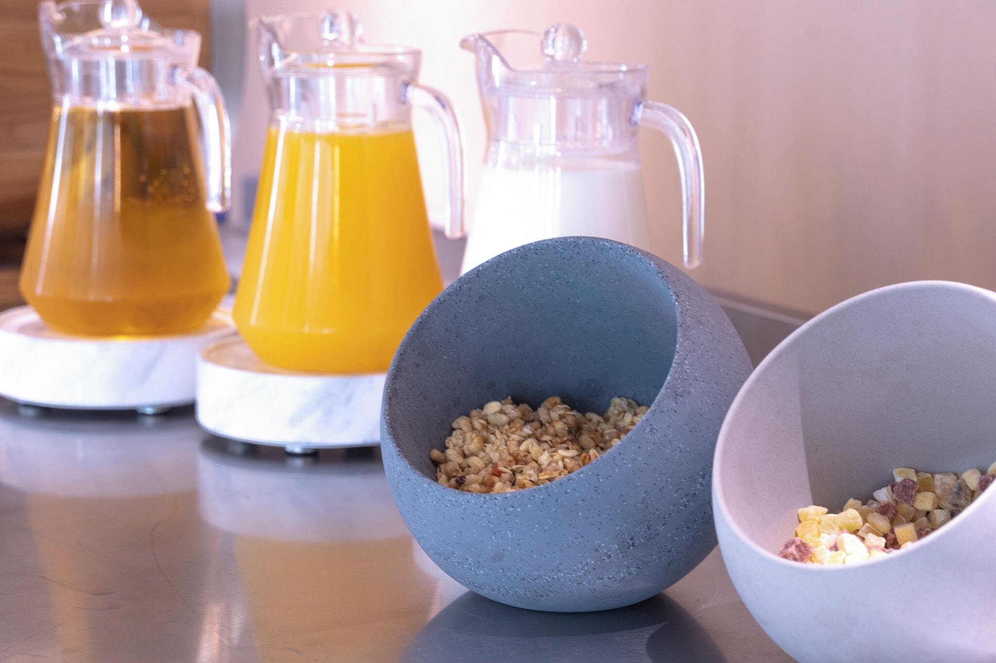 A kitchen counter with three pitchers of different liquids and two bowls containing grains and diced fruit.