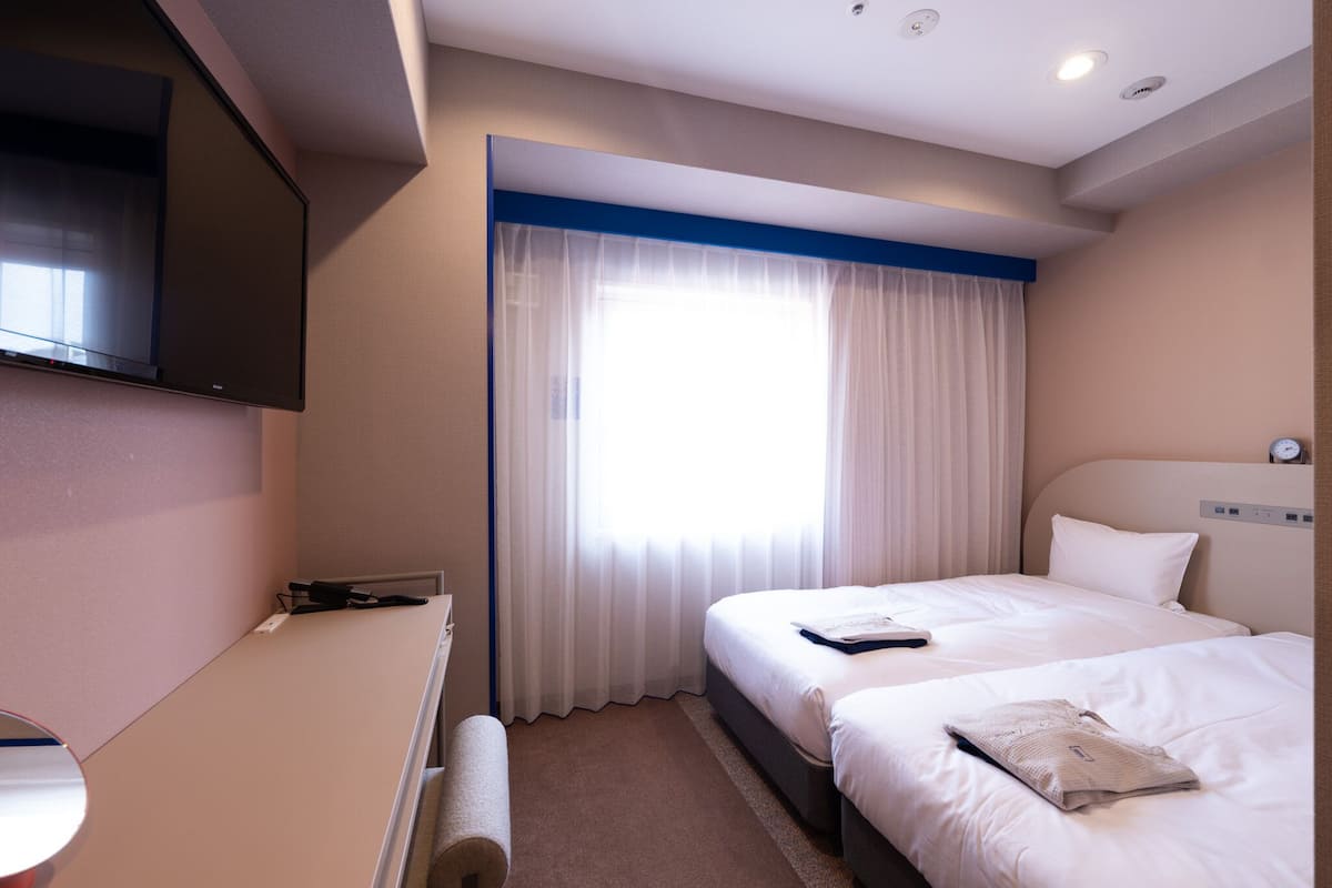 A hotel room with a flat-screen TV, two beds, a desk, and a window with curtains.