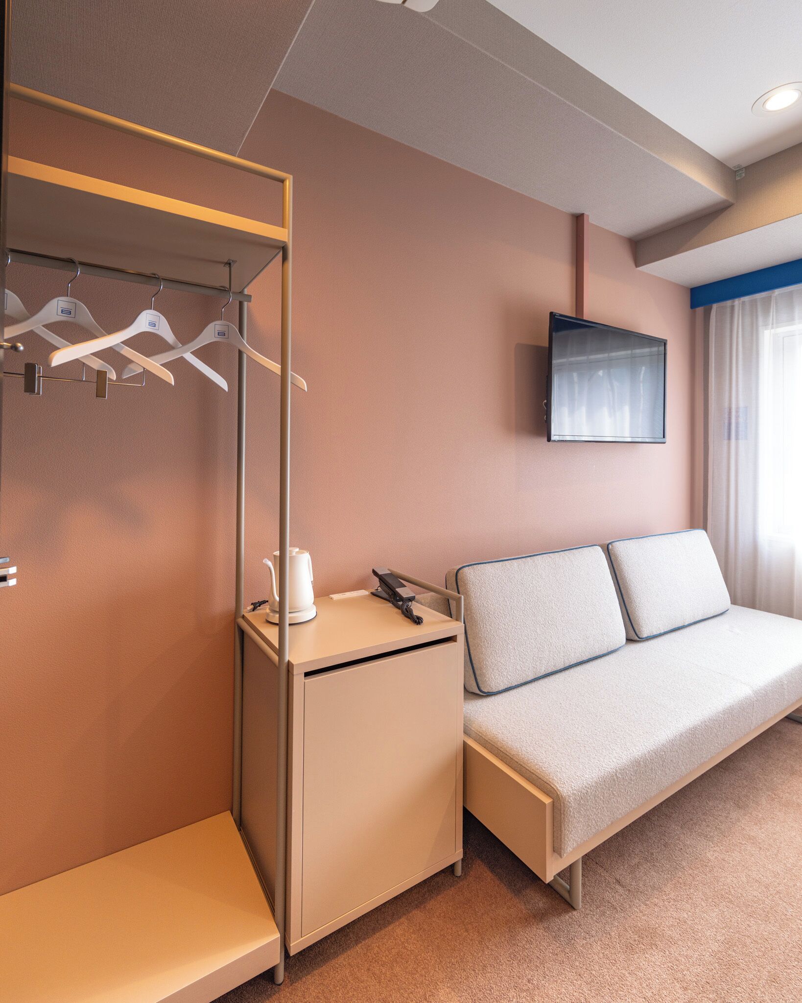 Una habitación de hotel con una cama, una mesita de noche pequeña con un teléfono y una tetera, un perchero y un televisor montado en la pared.
