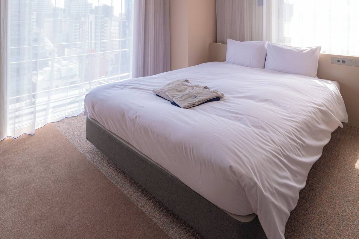 A hotel room with a large bed, two windows with sheer curtains, a city view, and a modern design.