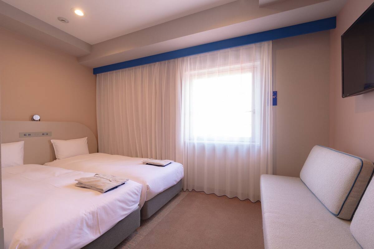 A hotel room with two beds, a flat-screen TV, a sofa, and a large window with sheer curtains.