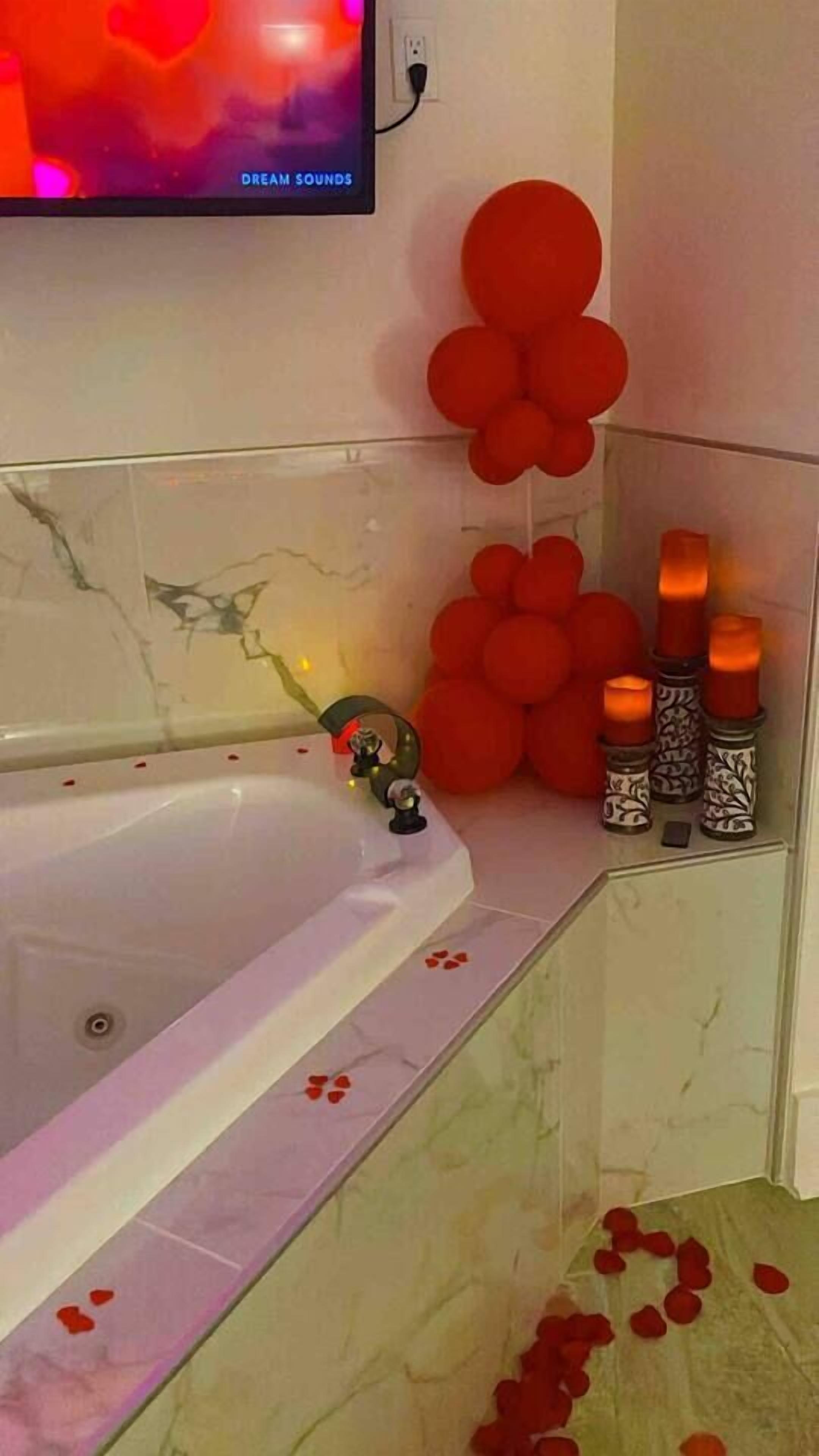 A bathroom with a bathtub, red balloons, candles, and rose petals on the floor.
