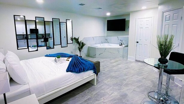 A modern hotel room with a bed, a sitting area with a glass table, a large mirror, a bathtub, and a TV mounted on the wall.