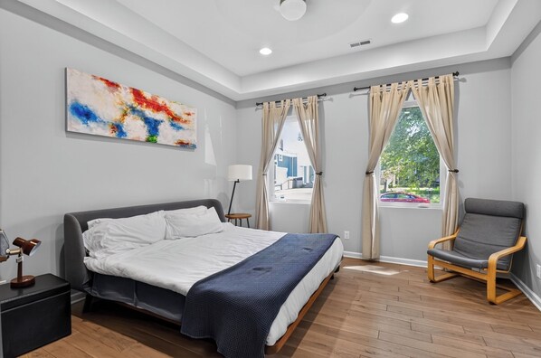 A modern bedroom with a large bed, a chair, a painting, and a window with curtains. - Entire Home in Fountain Square (Indianapolis)