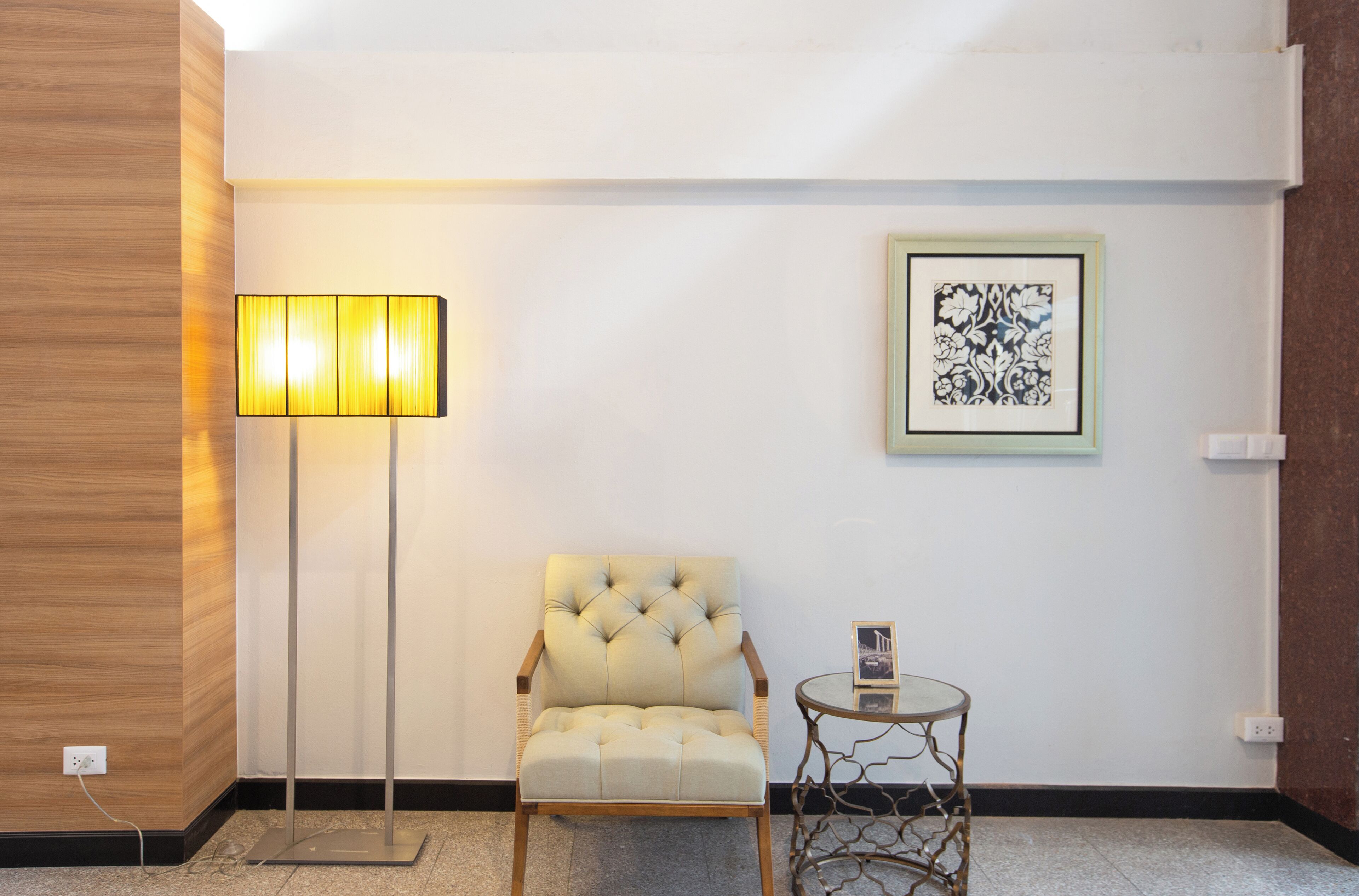 A room with a tufted armchair, a floor lamp, a small round table, and a framed artwork on the wall.