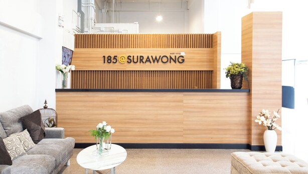 Un comptoir d'accueil avec un panneau décoratif en bois et l'inscription « 185@SURAWONG ». On y trouve un canapé, une petite table ronde avec un vase de fleurs et une plante en pot, le tout placé sur le côté droit.