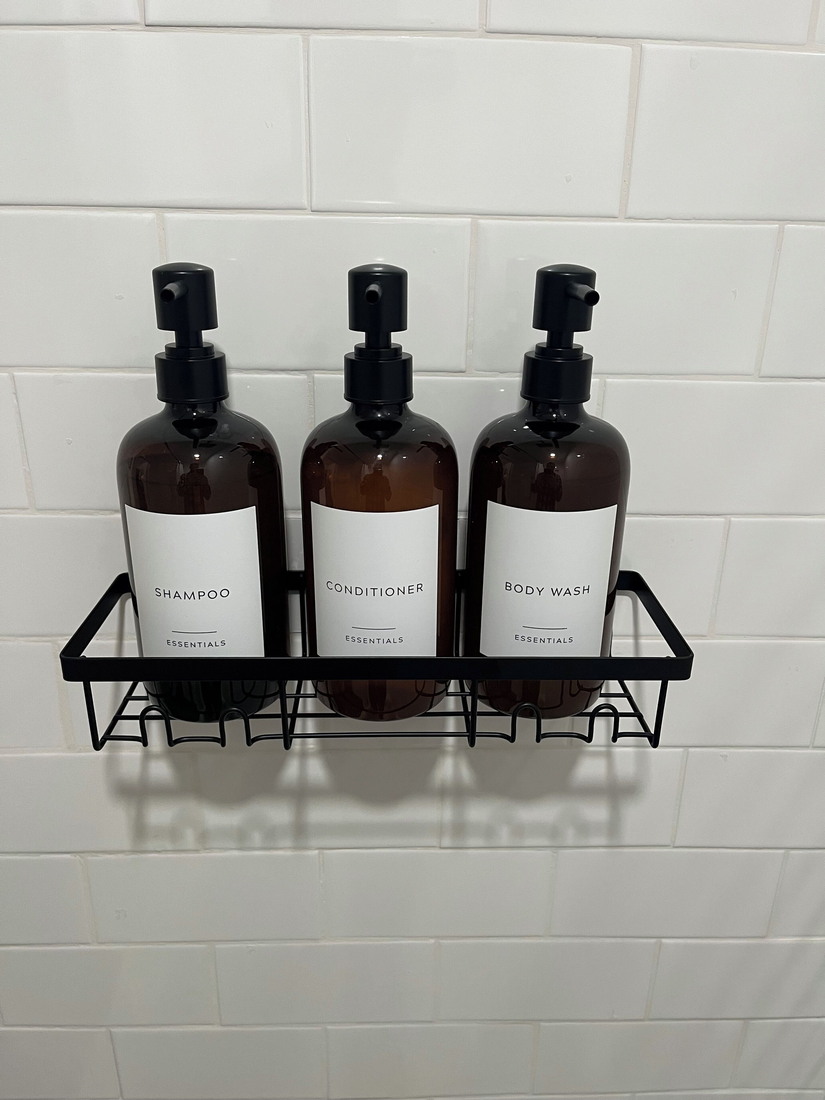 Three bottles of shampoo, conditioner, and body wash on a black wire shelf against a white tiled wall.