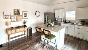 A modern kitchen with a central island, bar stools, a clock, and framed artwork on the wall. - Kit Carson Bungalow: AC/Heat + Pets Free! (Trinidad)