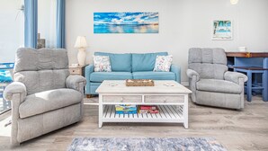 Un salon avec un canapé bleu, deux fauteuils gris, une table basse blanche et un tableau représentant une scène de plage.