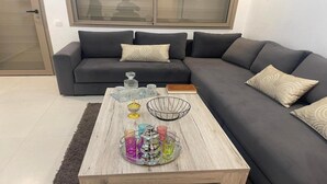 Un saló modern amb un sofà seccional gris fosc, una taula de centre de fusta i objectes decoratius, i una finestra gran amb persianes.