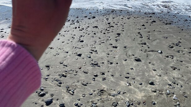 A hand holding a clear object near a beach with rocky terrain and a body of water in the background.