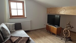 Une pièce comprenant un canapé, un téléviseur posé sur un meuble en bois, une fenêtre et un rouet.