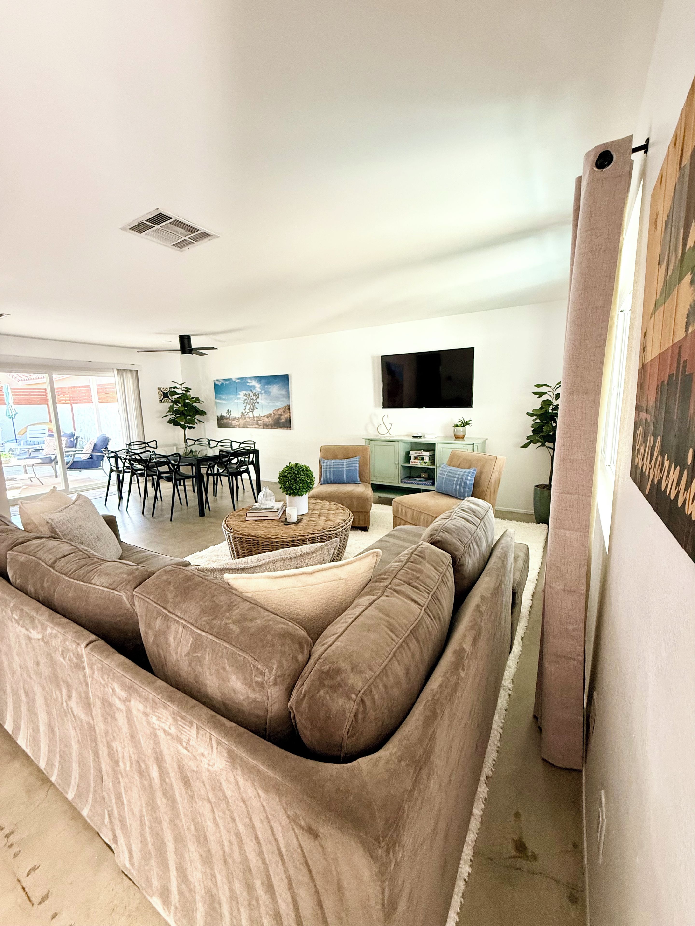 Ein Wohnzimmer mit einer großen, beigefarbenen Eckcouch, einem Essbereich mit Stühlen und Tisch, einem flachbildfernseher an der Wand und einem großen Fenster mit Blick ins Freie.