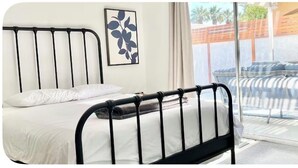 Un letto con struttura in metallo nero, materasso bianco e cuscino. C'è una sedia blu e una finestra che si affaccia su mobili da esterno.