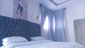 A neatly made bed with white linens in a room with a tufted headboard, a wall-mounted light fixture, and a window with curtains. - Luxury studio with Jacuzzi in a serene REstate (Lekki)