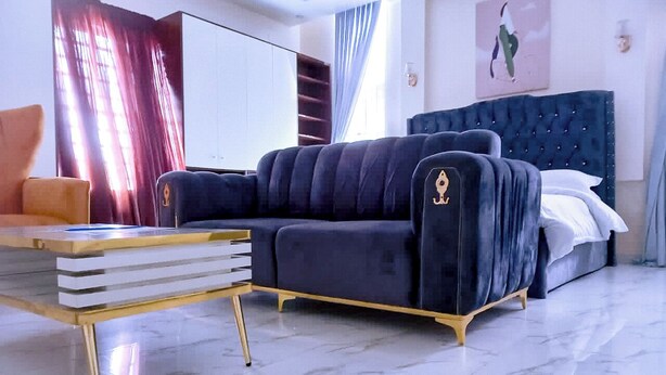 A modern bedroom with a dark blue upholstered sofa, a gold-framed coffee table, and a large bed with white linens. - Luxury studio with Jacuzzi in a serene REstate (Lekki)