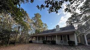 A single-story house with a porch, surrounded by trees and a leaf-covered ground. - Our Happy Place is located in the gated community of Holly Lake Ranch. (Hawkins)