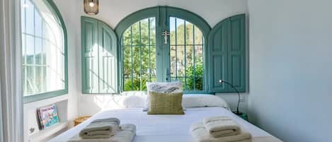A bedroom with a bed, two windows with green shutters, a bedside lamp, and a framed picture on the wall.