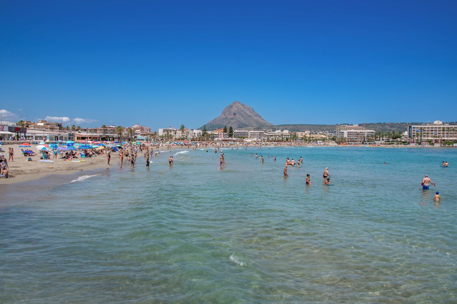 A beach with clear water, people swimming and sunbathing, and a mountain in the background.