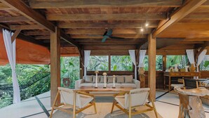 A wooden ceiling with a ceiling fan, a sofa with cushions, two wicker chairs, a wooden table with candles, and a large window with curtains. - Sungai Villa by Betterplace (Canggu)
