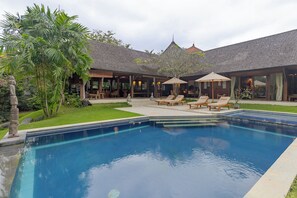 A swimming pool with lounge chairs and umbrellas, surrounded by a garden and a building with a tiled roof.