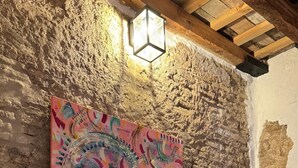 A rustic room with exposed brick walls, a wooden ceiling, a bed with white bedding, a colorful abstract painting, and a hanging light fixture.