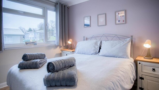A neatly made bed with pillows and rolled towels, a bedside table with a lamp, and a window with curtains.