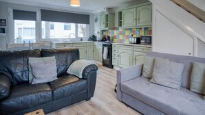 A cozy living room with a black leather sofa and a grey sofa, wooden flooring, and a kitchen with green cabinets and a colorful backsplash.