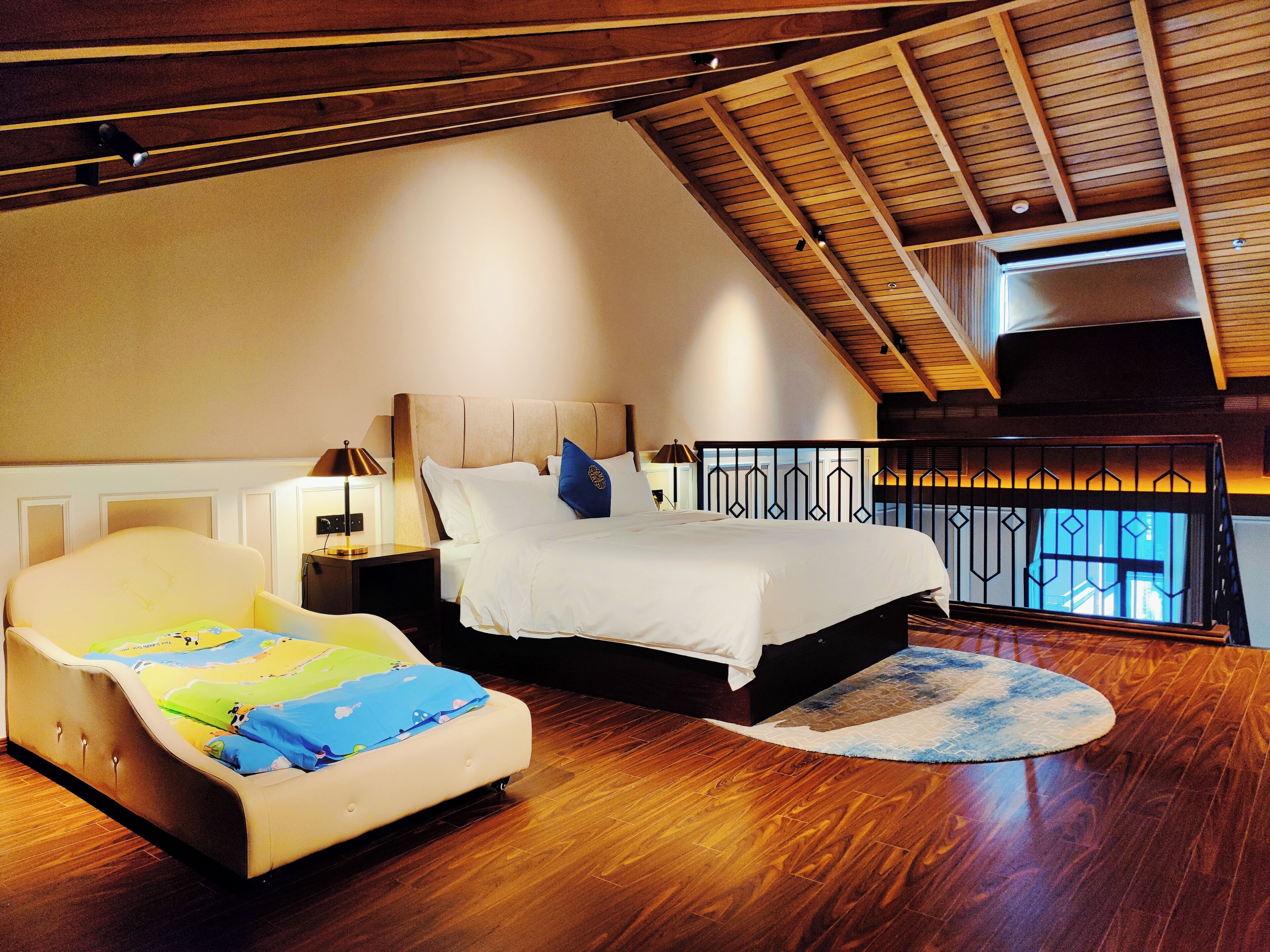A bedroom with a sloped ceiling, a bed with white bedding, a nightstand with a lamp, and a chair with a colorful blanket.