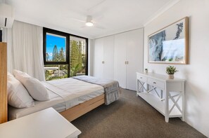 Quarto com cama, cómoda branca, ventoinha no teto e uma janela grande com vista para árvores e edifícios.