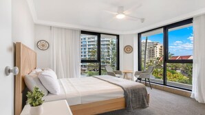 Una habitación de hotel moderna con una cama grande, una mesita pequeña, dos sillas, un ventilador de techo y grandes ventanales con vistas a la ciudad.