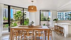 Un comedor moderno con una gran mesa de madera y sillas, una lámpara colgante de fibras tejidas y ventanas amplias que ofrecen vistas a un paisaje urbano y zonas verdes.